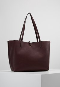 Borsa tote di un profondo bordeaux realizzata in pelle sintetica testurizzata, con due manici lunghi e un dettaglio metallico circolare nella parte superiore.