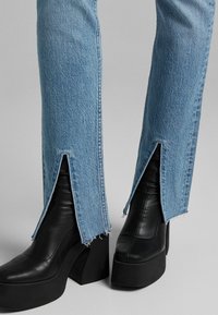 Helle blaue Jeans mit gespaltenem Saum, kombiniert mit schwarzen Plattform-Stiefeletten mit blockigem Absatz. Strukturiertes Material und rohen Kanten.
