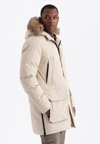 Béžová péřová bunda s kapucí vyloženou kožešinou, zapínáním na zip a knoflík, dvěma předními kapsami s klopami a kontrastními černými zipy. Měkká textura.