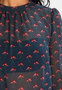 Blouse fleurie en tissu transparent bleu marine avec motifs de fleurs rouges, col froncé et texture douce. Léger avec des détails délicats.
