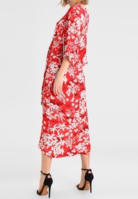 Robe fleurie rouge avec des motifs blancs, coupe ample, manches trois-quarts, longueur genou, tissu léger, portée avec des sandales à talons noirs.