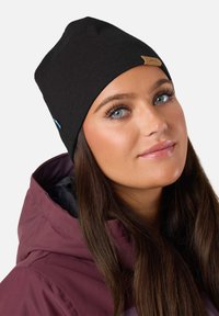 Czarna czapka beanie z miękkiego materiału, z jasnobrązowym skórzanym logo. Noszona z bordową kurtką z kapturem.