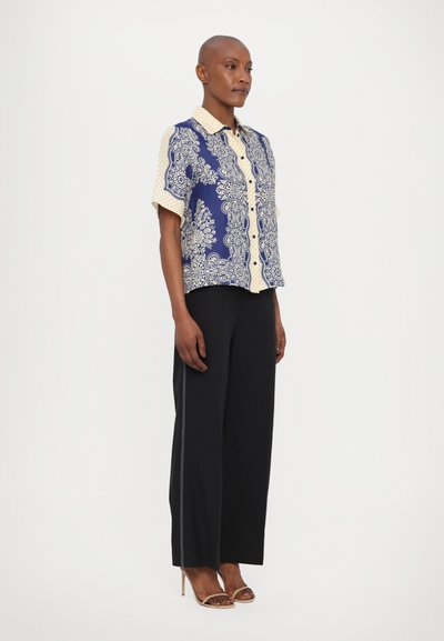 Blusa a maniche corte con un motivo paisley blu e crema, dotata di una chiusura a bottoni frontale e un design con colletto. Indossata con pantaloni neri.