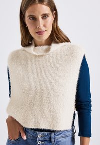 Pull en tricot de couleur crème avec un col montant, présentant une finition douce et texturée et des côtés ouverts, superposé sur un haut à manches longues bleu.