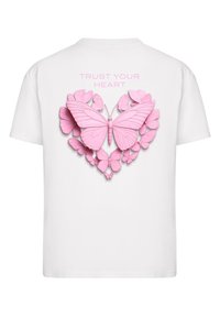 Weißes T-Shirt mit einem 3D-Design von rosa Schmetterlingen, die in Herzform angeordnet sind, mit dem Text "VERTRAUE DEINEM HERZEN" darüber.