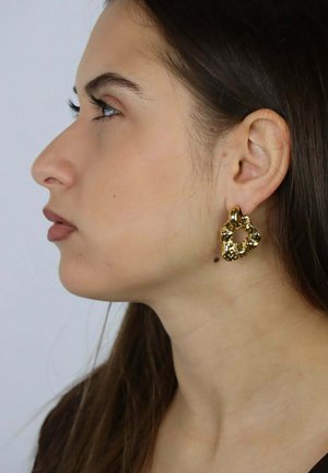 Pendientes de aro de oro con textura que presentan una forma orgánica e irregular. Lucidos por una modelo sobre un fondo neutral, destacando su diseño.