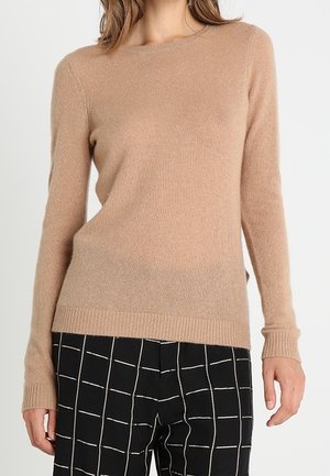 Frau trägt einen beige taillierten Langarmpullover kombiniert mit schwarzen Hosen mit weißem Gittermuster.