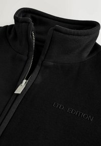 Black zip-up sweatshirt with a high collar, featuring a smooth texture, silver zipper, and embossed "LTD. EDITION" text on the front.