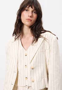 Cremefarbener gestreifter Anzug mit Blazer und Weste. Der Stoff hat eine leichte Textur. Der Blazer verfügt über strukturierte Revers und drei Knopfverschlüsse.