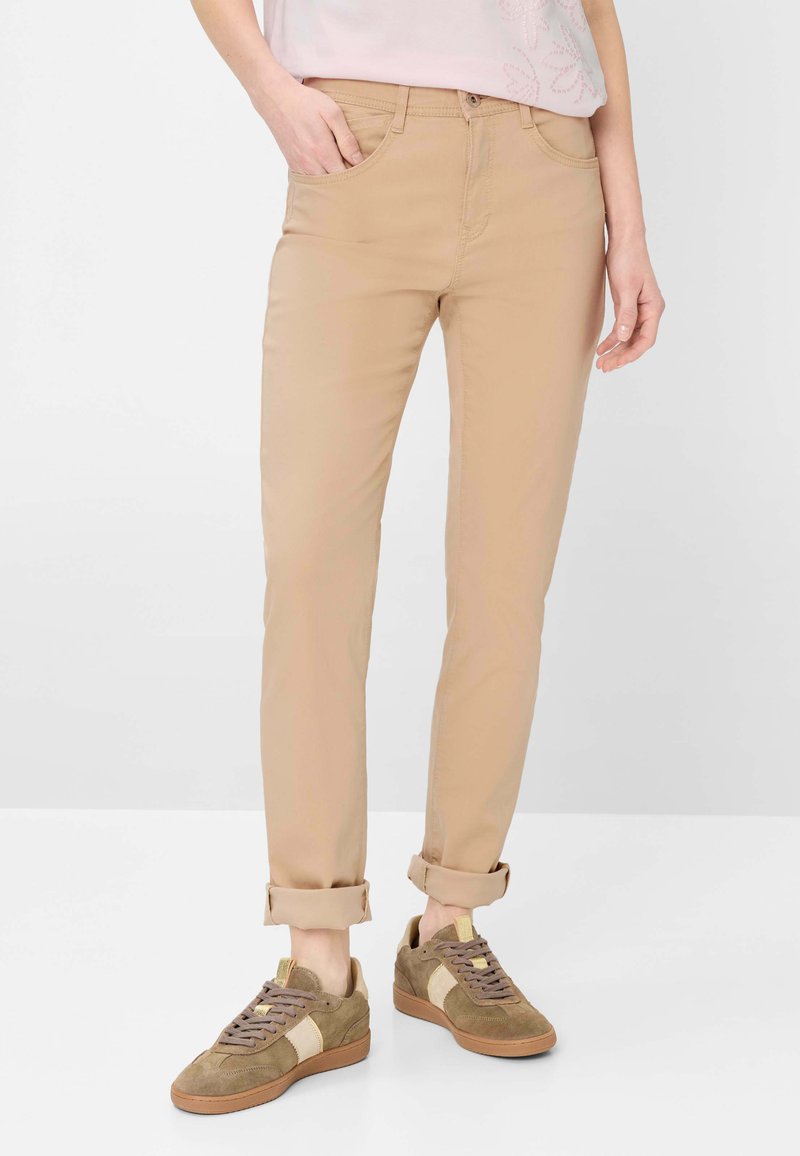 Person trägt beige, schmal geschnittene Hose, die am Saum umgekrempelt ist, und braune Wildleder-Sneaker mit einem hellen Streifen.
