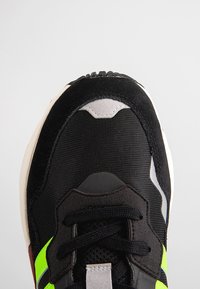 Svart sneakers i mesh och mocka, med en grön accentrand, texturerade snören och en vit sula. Rundad tådesign.