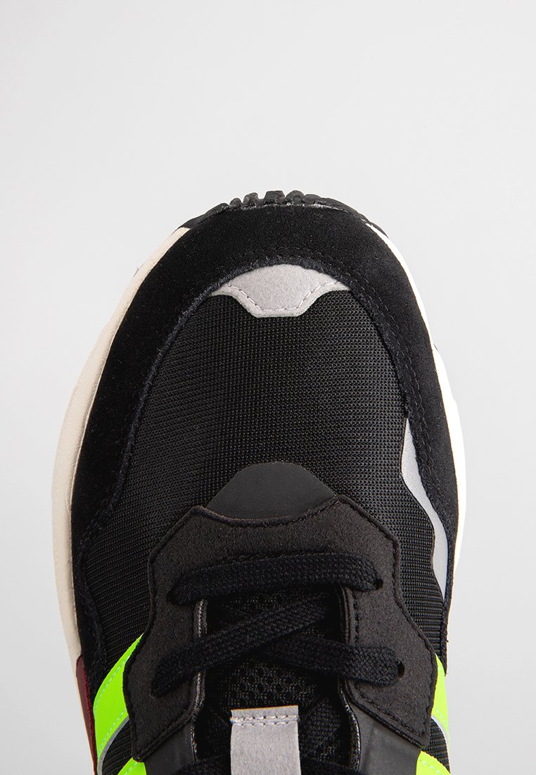 Svart sneakers i mesh och mocka, med en grön accentrand, texturerade snören och en vit sula. Rundad tådesign.