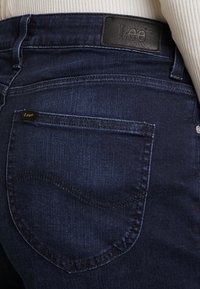 Tmavě modré džíny z džínoviny mají vysoký pas, šité kapsy a koženou nálepku se značkou "Lee" vzadu. Hebká textura a klasický střih.