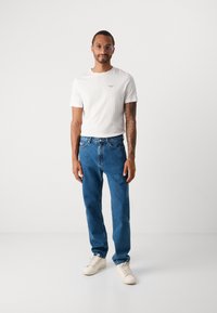 Mann som bruker briller, hvit t-skjorte, blå jeans og hvite joggesko som står mot en enkel hvit bakgrunn.