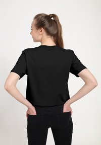 Černé tričko s krátkým rukávem a kulatým výstřihem, s jednoduchým designem. Model má na sobě černé džíny a stojí s rukama v kapsách.