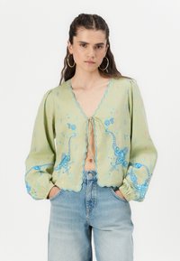 Ljusgrön blus med scallopkanter, blå blommig broderi och voluminösa ärmar. Har en knytning vid halsringningen.