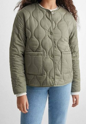 Personne portant une veste matelassée vert olive clair avec deux poches avant, des boutons-pression et un jean bleu clair.