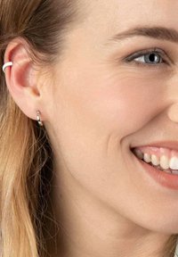 Silberne Creolen mit glatter Oberfläche, die am Ohrläppchen getragen werden, kombiniert mit einem dezenten weißen Ohrbänder. Helle Haut und glattes Haar sind sichtbar.