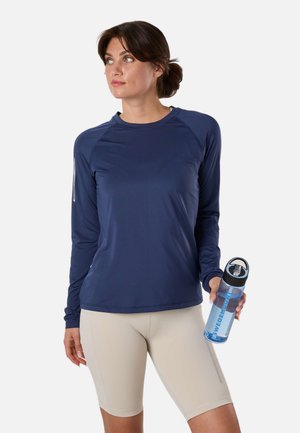 Kvinde i navyblå langærmet træningstop og beige shorts, der holder en klar vandflaske og kigger mod venstre med håret samlet bagud.