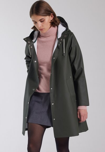 Olijfgroen waterdichte jas met een witte capuchonvoering, drukknopen en zijzakken, gedragen over een lichtroze trui en een donkergrijze rok.