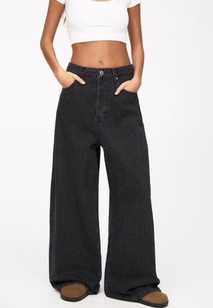 Woman wearing high-waist, wide-leg black jeans with hands in pockets and brown shoes, paired with a white cropped short-sleeve top.