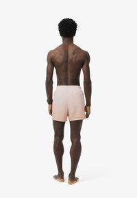 Costume da bagno rosa chiaro con cintura elastica, tasca posteriore e finitura in tessuto liscia, indossato da una persona vista di spalle.