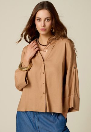 Jonge vrouw met lang bruin haar die een losse beige blouse met knopen, blauwe broek, gouden armband en laagjes kettingen draagt, kijkt recht vooruit.