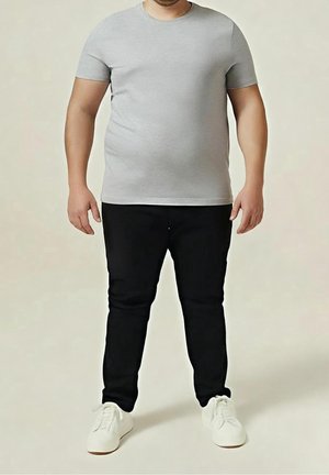 Mężczyzna ubrany w prostą szarą koszulkę z krótkim rękawem, czarne spodnie i białe tenisówki, stojący na neutralnym tle.