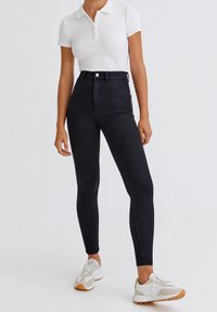 Jeans skinny negros de cintura alta con una textura suave y diseño de cinco bolsillos, combinados con una camiseta polo blanca y zapatillas blancas.