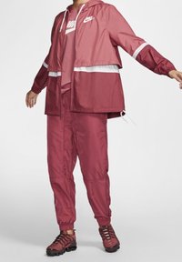Ensemble de sport Nike rouge comprenant une veste à capuche avec des accents blancs et un pantalon assorti. Le tissu semble léger et lisse.