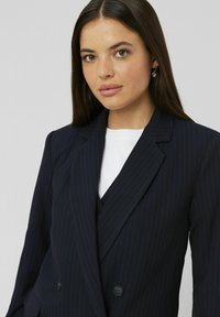 Blazer bleu marine à rayures fines avec un design à double boutonnage, des revers à cran et un tissu lisse. Stylé avec un top blanc en dessous.
