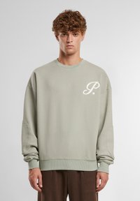 Lys grøn genser med rund hals, senket skuldre, ribbestrikkede mansjetter og en hvit brodert logo på venstre bryst. Myk tekstur.
