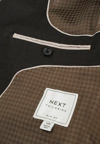 Schmal geschnittene Tailored-Anzugjacke in Dunkelbraun mit einem strukturierten Hahnentritt-Muster, kontrastierender sandfarbener Futter und einem einzelnen schwarzen Knopfverschluss.