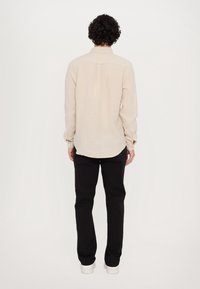 Uomo con capelli ricci che indossa una camicia a maniche lunghe beige, pantaloni neri e sneakers bianche, in piedi con le spalle alla macchina fotografica.