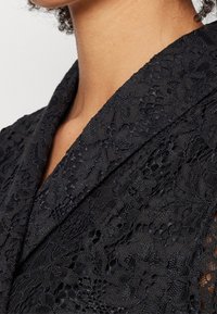 Gros plan d'une personne portant un blazer en dentelle florale noire avec un col à revers cranté sur un fond clair.