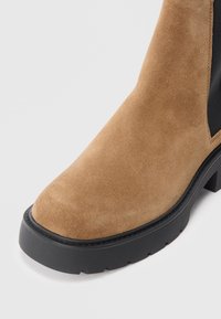 Brown suede ankle boot with a rounded toe, elastic side panels, and a black rubber sole featuring a treaded pattern.