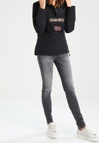 Czarny sweter z zamkiem, z kieszenią na piersi i naszywką z norweską flagą. W zestawieniu z szarymi dżinsami i białymi trampkami.