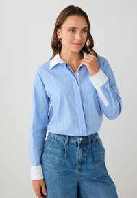 Vrouw in een lichtblauwe gestreepte blouse met witte kraag en manchetten, gecombineerd met hoge blauwe jeans, staand tegen een neutrale achtergrond.