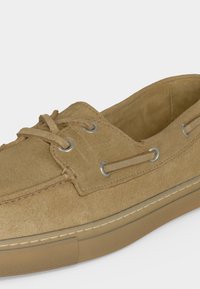 Ανοιχτό καφέ παπούτσι τύπου boat από σουέτ με δερμάτινα κορδόνια περασμένα μέσα από μεταλλικά κορδόνια και ελαστική σόλα με λευκό ράψιμο.