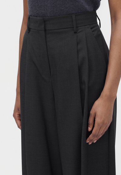 Pantalon noir taille haute, ajusté, avec plis, passants pour ceinture et poches latérales, porté avec un haut gris foncé.