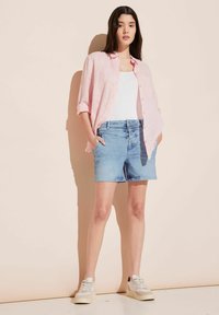 Camisa de lino rosa claro sobre una camiseta blanca, pantalones cortos de mezclilla azules con bolsillos delanteros, y zapatillas blancas con acentos beige.
