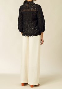 Blusa in pizzo nero con ritagli decorativi, colletto alto e maniche a sbuffo, abbinata a pantaloni larghi color crema e sandali con tacco block arancioni.