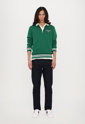 Pullover verde con strisce bianche, colletto con zip e logo "POLO SPORT", abbinato a jeans scuri e sneaker bianche. Design semplice, vestibilità casual.