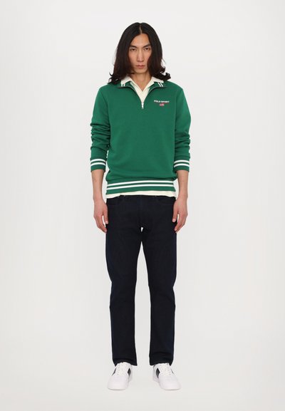 Grønn genser med hvite striper, glidelås ved kragen, og "POLO SPORT" logo, kombinert med mørke jeans og hvite sneakers. Enkelt design, uformell passform.