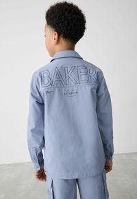 Svetlomodrá dlhá košeľa s dlhým rukávom s vyšitým textom „BAKER“ na chrbte, s gombíkmi na rukávoch a vo voľnom strihu. Hladká tkanina.