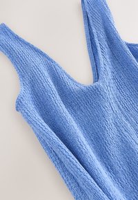 Débardeur en maille texturée bleue avec larges bretelles et motif ondulé subtil, posé à plat sur une surface claire.