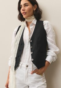 Črna lanena jopica s petimi gumbi, kombinirana z belo srajco in belimi hlačami. Obleko dopolnjuje lahka ruta z čipkastimi detajli.