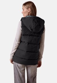 Schwarze wattierte Weste mit Kapuze, mit horizontaler Steppung, weicher Textur und getragen über einem hellgrauen Langarmoberteil.