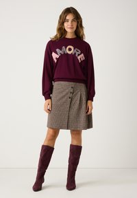Frau in einem burgunderroten Pullover mit dem Text "AMORE", einem knopfbaren Tweedrock und passenden burgunderbraunen Wildlederstiefeln bis zum Knie, steht vor einer weißen Wand.