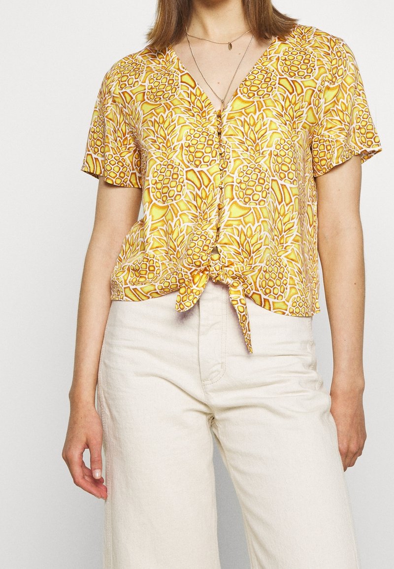 Femme portant une chemise jaune à manches courtes avec motif ananas, nouée à la taille, et un pantalon taille haute blanc cassé.